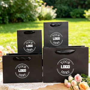 May include: Four black paper shopping bags of varying sizes, each featuring black ribbon handles and a circular white graphic with the text YOUR LOGO HERE centered on the front. The bags are arranged in a group on a white lace-covered surface.