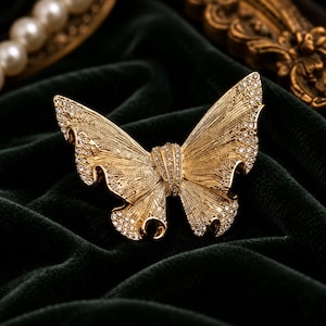 May include: A gold-toned butterfly brooch featuring a textured, ribbed metal surface and small, clear, sparkling crystals embedded along the edges of the wings and the central body section of the insect-shaped accessory.