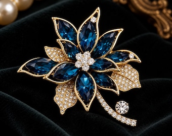 May include: A gold-tone flower brooch featuring deep blue marquise-cut crystals as petals, a central cluster of small clear round crystals, and two gold leaves encrusted with tiny clear stones, with a crystal-accented stem.