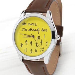 May include: A silver wristwatch with a yellow face and a brown leather band. The watch face has black handwritten text that reads "who cares, I'm already late." The watch hands point to approximately 10:08. The band is approximately 0.75 inches wide.