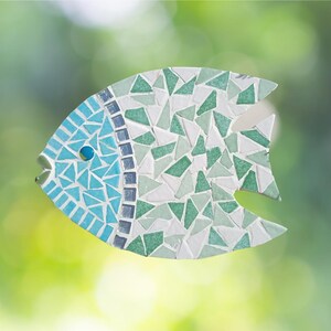 May include: A mosaic fish sculpture with a blue, green, and white color scheme. The fish has a blue eye and is made of small, irregularly shaped tiles. The background is a blurred green.