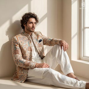 May include: A beige bandhgala jacket featuring a multi-colored geometric and floral pattern in shades of blue, orange, and brown, worn over a white kurta and white trousers. The jacket has a mandarin collar and a dark blue pocket square.