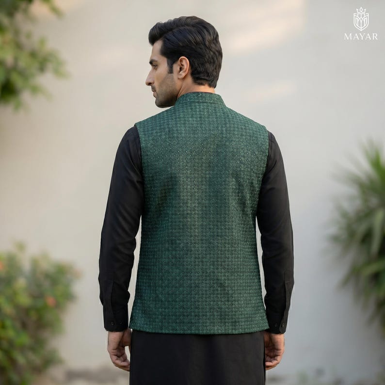 May include: A dark green patterned vest with a mandarin collar, worn over a black long-sleeved shirt and black pants. The vest has a textured, woven appearance. The person is seen from the back, with a neutral background.