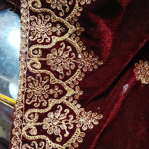 May include: A deep burgundy velvet fabric featuring an intricate border of metallic gold embroidery. The design consists of repeating floral motifs enclosed within scalloped arches, with additional small floral patterns scattered across the velvet.