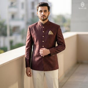 May include: A dark maroon bandhgala jacket with a subtle textured pattern, featuring a high collar, a row of five gold-toned buttons, and a light-coloured pocket square in the chest pocket, paired with plain off-white trousers.