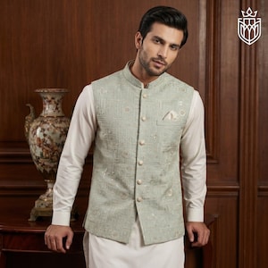 May include: A light green vest with an intricate geometric pattern and a mandarin collar. The vest has a pocket on the chest and is buttoned up. The vest is worn over a white long-sleeved shirt and white pants.