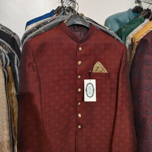 May include: A deep maroon bandhgala jacket featuring a subtle repeating geometric pattern, a mandarin collar, and a front row of gold-toned buttons. A gold pocket square is tucked into the chest pocket, and a white tag labelled Mayar is attached.