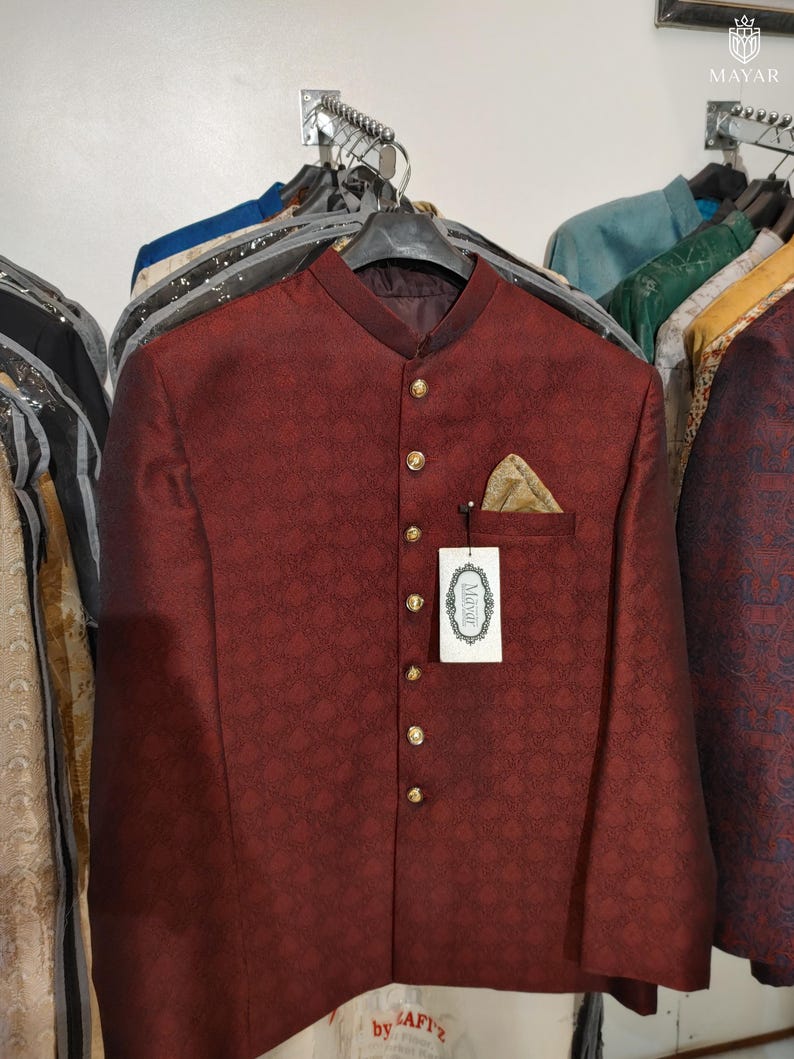 May include: A deep maroon bandhgala jacket featuring a subtle repeating geometric pattern, a mandarin collar, and a front row of gold-toned buttons. A gold pocket square is tucked into the chest pocket, and a white tag labelled Mayar is attached.