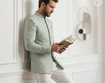 May include: A light green jacket with a mandarin collar and intricate embroidery, paired with white pants. The jacket has gold buttons and a pocket square. The person is holding a book, leaning against a white wall.