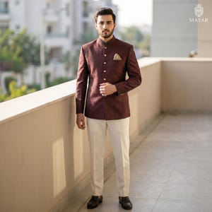 May include: A dark maroon bandhgala jacket with a high collar and a row of gold buttons, paired with cream-coloured trousers. The jacket features a gold pocket square in the chest pocket. The ensemble is presented with the Mayar brand logo visible.