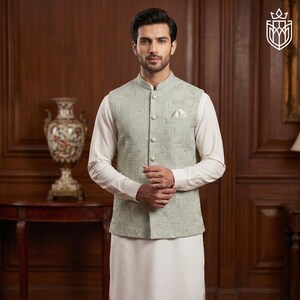 May include: A light green embroidered vest with a mandarin collar, worn over a white long-sleeved shirt and white pants. The vest has a geometric pattern and is buttoned up. A pocket square is visible. The outfit is formal wear.