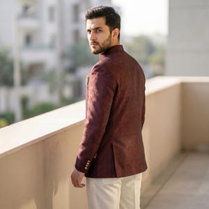 May include: A dark maroon blazer with a subtle textured finish, featuring a high collar, long sleeves with four small buttons at the cuffs, and a single pocket square detail on the chest, paired with plain off-white trousers.