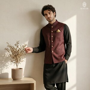 May include: A dark maroon Nehru jacket with a subtle textured pattern, featuring a row of gold-toned buttons and a gold pocket square. The jacket is worn over a solid black long-sleeved shirt and matching black trousers, creating a formal ensemble.