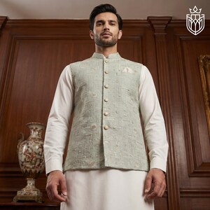 May include: A light green embroidered vest with a mandarin collar and gold buttons. The vest is worn over a white long-sleeved shirt and white pants. The vest features a detailed geometric pattern.