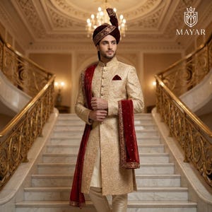 May include: A beige sherwani with intricate gold embroidery, featuring a long-sleeved jacket, matching trousers, and a deep red velvet stole. The ensemble includes a matching maroon turban with gold detailing and a pocket square. Text reads MAYAR.