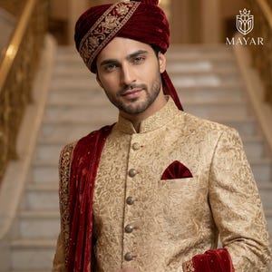 May include: A beige sherwani featuring an intricate tonal floral pattern, a mandarin collar, and a row of metallic buttons. A deep red velvet pocket square and a matching red velvet turban with gold embroidery are worn with a red velvet stole.
