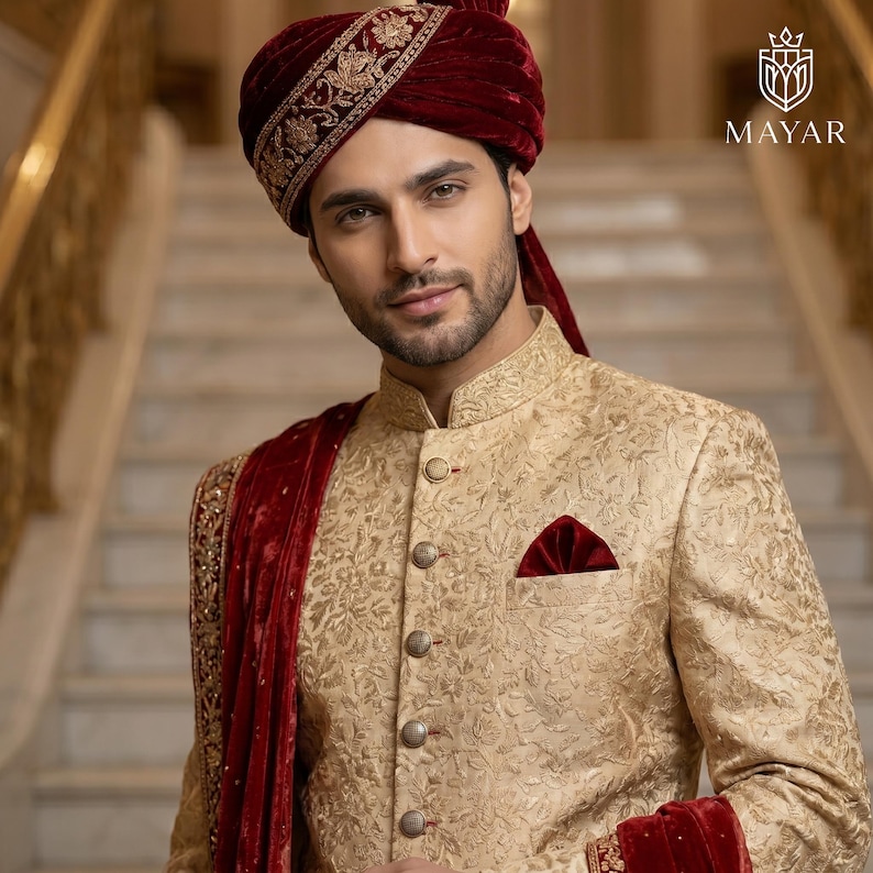 May include: A beige sherwani featuring an intricate tonal floral pattern, a mandarin collar, and a row of metallic buttons. A deep red velvet pocket square and a matching red velvet turban with gold embroidery are worn with a red velvet stole.