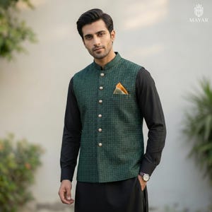 May include: A green patterned vest with gold buttons and a pocket square. The vest is worn over a black long-sleeve shirt and black pants. A watch is visible on the wrist.