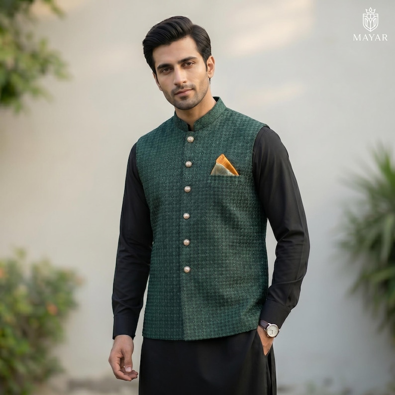 May include: A green patterned vest with gold buttons and a pocket square. The vest is worn over a black long-sleeve shirt and black pants. A watch is visible on the wrist.