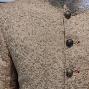 May include: A beige mandarin collar jacket featuring intricate tonal floral embroidery throughout the fabric. The garment is fastened with four round, textured metallic buttons, each secured with a small red thread detail at the buttonhole.