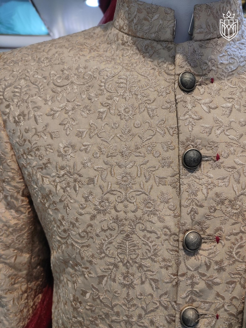 May include: A beige mandarin collar jacket featuring intricate tonal floral embroidery throughout the fabric. The garment is fastened with four round, textured metallic buttons, each secured with a small red thread detail at the buttonhole.