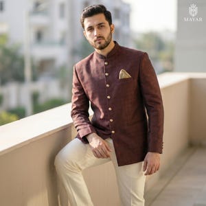 May include: A dark maroon bandhgala jacket featuring a mandarin collar, a single row of metallic buttons, and a tan pocket square, paired with off-white trousers. The jacket has a subtle textured pattern and long sleeves with buttoned cuffs.