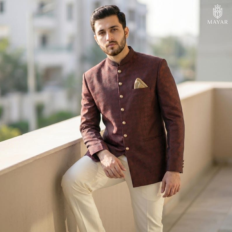 May include: A dark maroon bandhgala jacket featuring a mandarin collar, a single row of metallic buttons, and a tan pocket square, paired with off-white trousers. The jacket has a subtle textured pattern and long sleeves with buttoned cuffs.