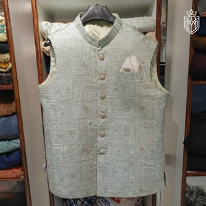 May include: A light green vest with a textured, geometric pattern. The vest has a mandarin collar, a pocket square, and six buttons down the front. The vest is sleeveless and appears to be made of a woven fabric.