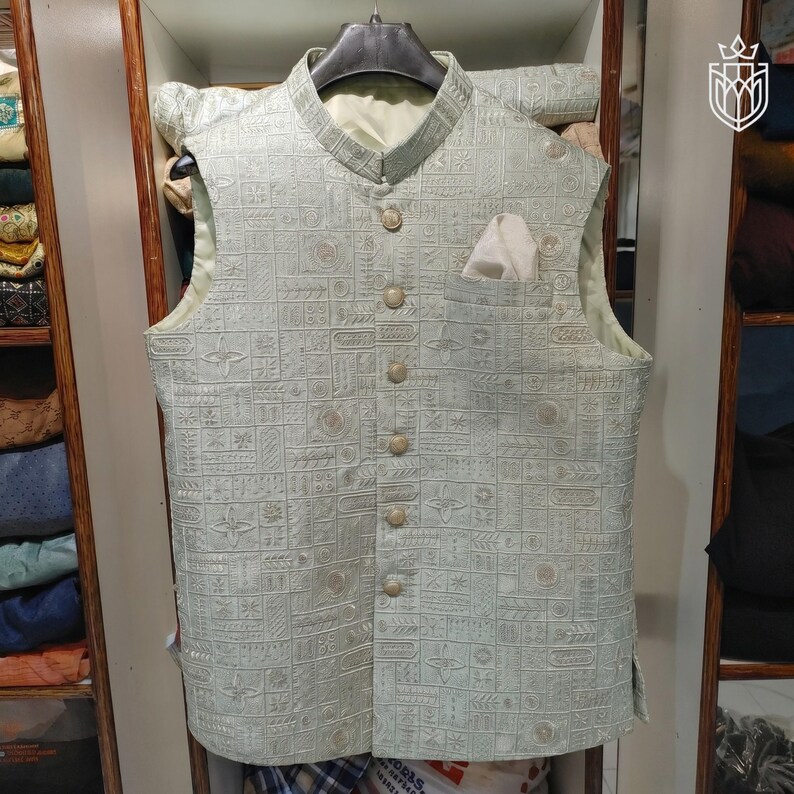 May include: A light green vest with a textured, geometric pattern. The vest has a mandarin collar, a pocket square, and six buttons down the front. The vest is sleeveless and appears to be made of a woven fabric.