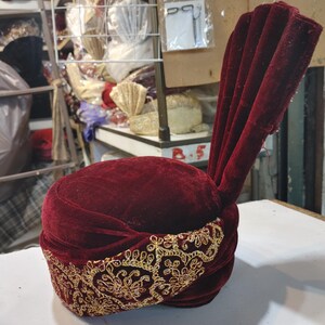 May include: A deep burgundy velvet turban featuring a prominent, upright pleated fan detail on one side. The base of the headpiece is wrapped in a wide band of intricate gold metallic embroidery, showcasing a repeating floral and scrollwork pattern.