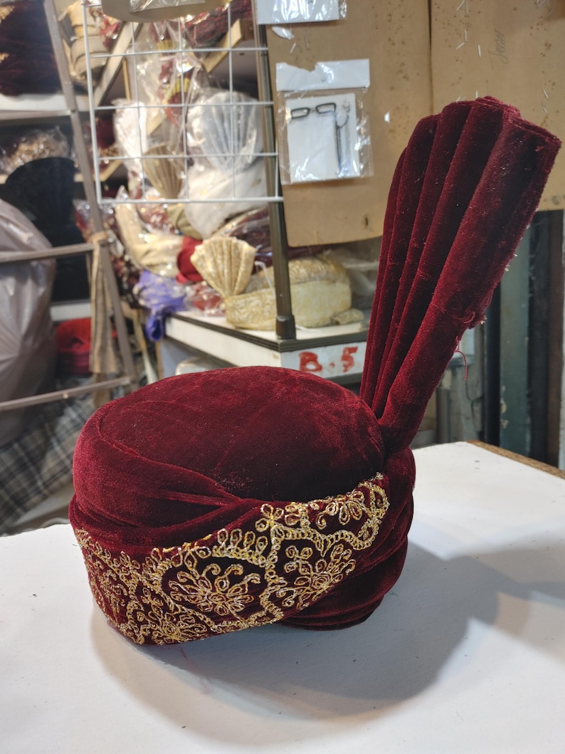May include: A deep burgundy velvet turban featuring a prominent, upright pleated fan detail on one side. The base of the headpiece is wrapped in a wide band of intricate gold metallic embroidery, showcasing a repeating floral and scrollwork pattern.