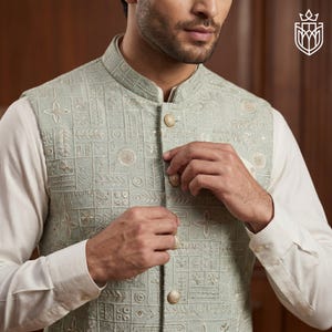 May include: A light green embroidered vest with a mandarin collar and gold buttons. The vest features a geometric pattern with floral and circular designs. The vest is worn over a white long-sleeved shirt.