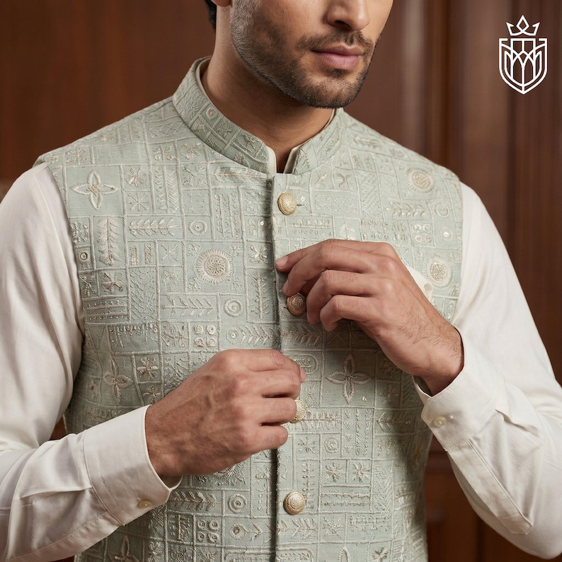 May include: A light green embroidered vest with a mandarin collar and gold buttons. The vest features a geometric pattern with floral and circular designs. The vest is worn over a white long-sleeved shirt.