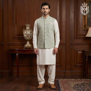 May include: A man wearing a light green vest with a subtle pattern over a white long-sleeved shirt and white pants. The vest has a mandarin collar and button closure. The outfit is paired with brown sandals.