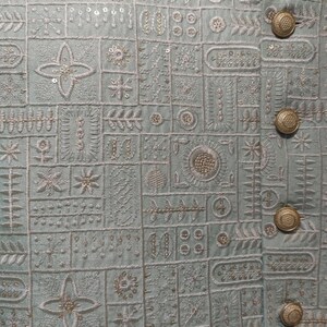 May include: Close-up of a light blue jacket with intricate embroidered patterns. The jacket features a series of geometric and floral designs in white thread, with gold-colored buttons. The overall aesthetic is detailed and elegant.