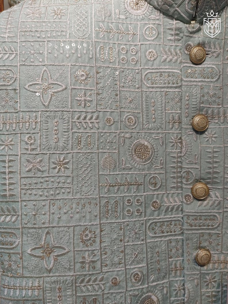 May include: Close-up of a light blue jacket with intricate embroidered patterns. The jacket features a series of geometric and floral designs in white thread, with gold-colored buttons. The overall aesthetic is detailed and elegant.