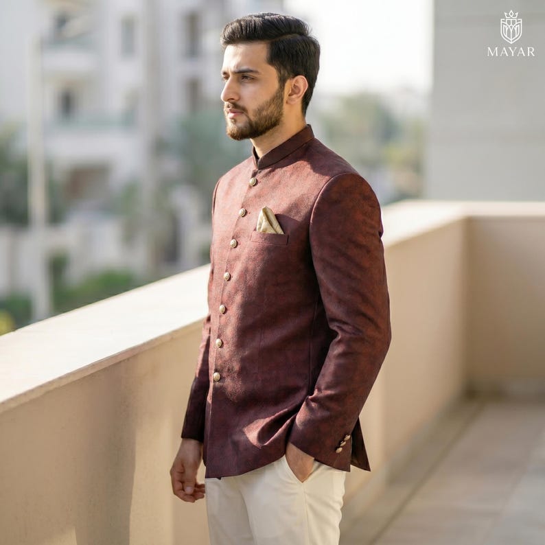 May include: A dark maroon bandhgala jacket featuring a high collar, long sleeves, and a row of metallic buttons down the front. The jacket has a textured fabric finish and a small pocket square tucked into the chest pocket, paired with white trousers.