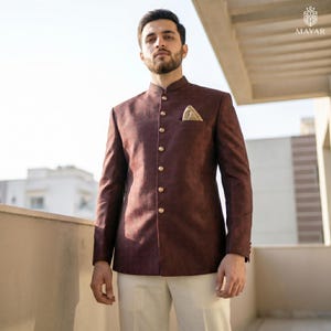 May include: A dark maroon bandhgala jacket featuring a subtle textured pattern, a mandarin collar, and a row of gold-toned buttons down the front. A small gold pocket square is tucked into the chest pocket, paired with plain cream trousers.