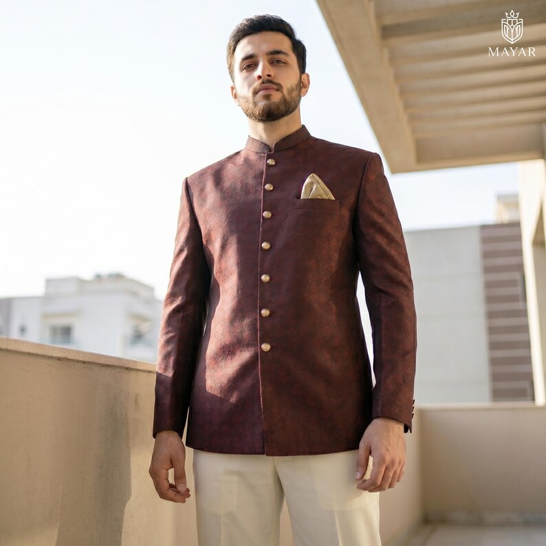 May include: A dark maroon bandhgala jacket featuring a subtle textured pattern, a mandarin collar, and a row of gold-toned buttons down the front. A small gold pocket square is tucked into the chest pocket, paired with plain cream trousers.