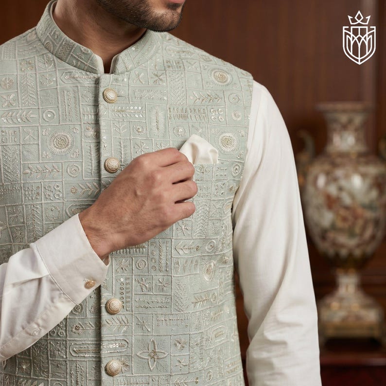 May include: A light green embroidered vest with a geometric pattern and gold buttons. The vest is worn over a white long-sleeve shirt. A white pocket square is in the vest pocket. The vest has a mandarin collar.