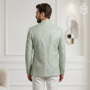 K&ouml;nnte beinhalten: Hellgr&uuml;ner Blazer mit dezentem geometrischem Muster. Der Blazer hat einen Stehkragen und goldfarbene Kn&ouml;pfe. Die Person tr&auml;gt eine wei&szlig;e Hose. Der Blazer ist ein formeller Stil.