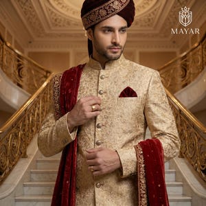 May include: A beige sherwani featuring intricate gold embroidery, a mandarin collar, and metallic buttons. A deep red velvet stole with gold trim is draped over the shoulder. The ensemble includes a matching red velvet turban with gold detailing.