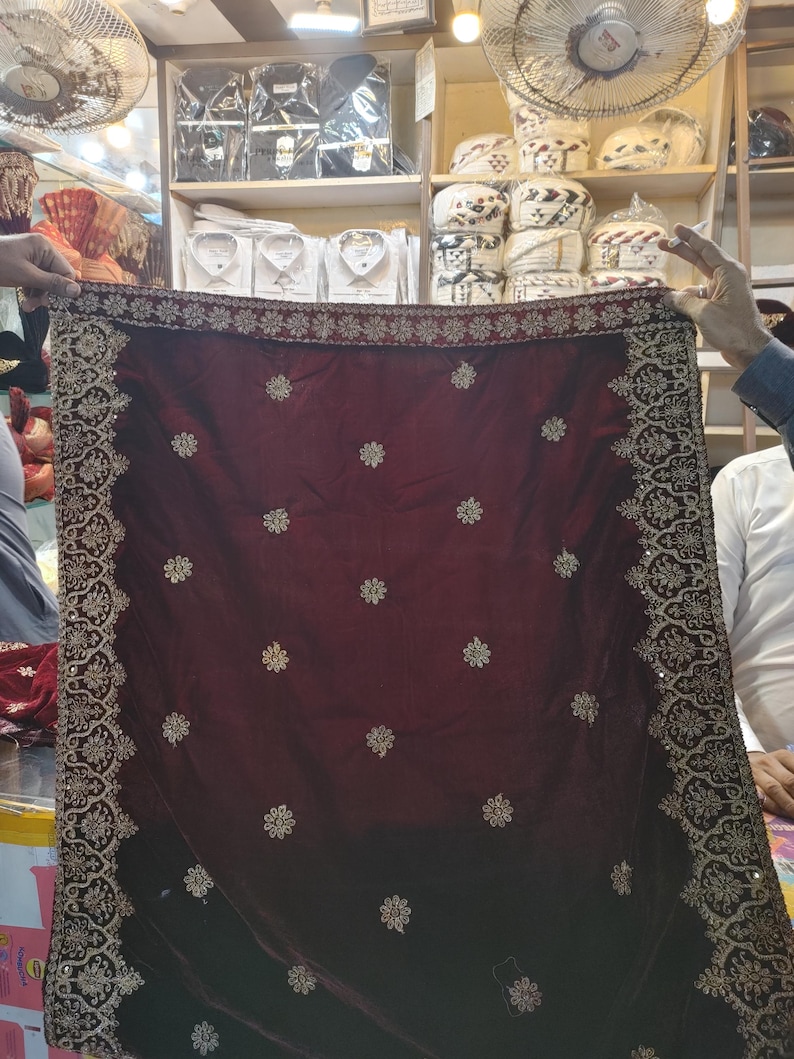 May include: A deep maroon velvet shawl featuring an intricate gold embroidered border and scattered gold floral motifs across the fabric. The rectangular textile displays a dense, ornate pattern along all four edges with small, repeating flower designs.