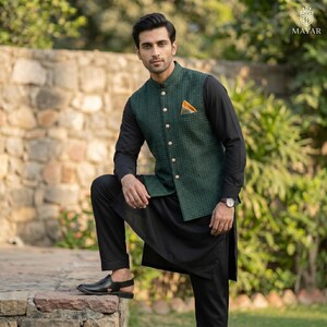 May include: A man wearing a green patterned vest with gold buttons, a black long-sleeve shirt, and black pants. The vest has a pocket square. The outfit is completed with black sandals and a watch. The background features a stone wall and greenery.