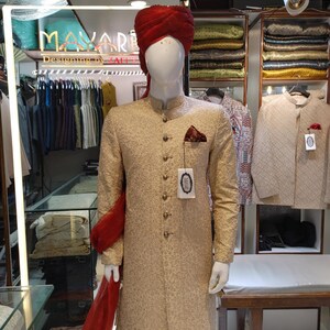 May include: A beige sherwani with a textured pattern, featuring a mandarin collar, long sleeves, and a row of metallic buttons. A deep red turban and a matching red fabric drape are worn with the garment. A pocket square is tucked into the chest.