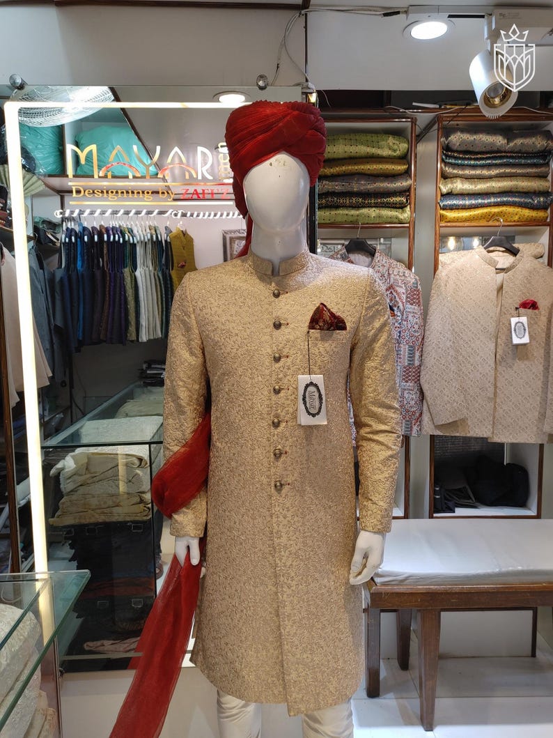 May include: A beige sherwani with a textured pattern, featuring a mandarin collar, long sleeves, and a row of metallic buttons. A deep red turban and a matching red fabric drape are worn with the garment. A pocket square is tucked into the chest.