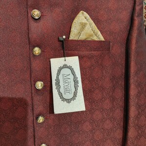 May include: A deep maroon jacket with a subtle repeating geometric pattern, featuring gold-toned buttons and a gold pocket square. A rectangular white tag hangs from the pocket, displaying the text Mayar The Trend Setter Designer Boutique.