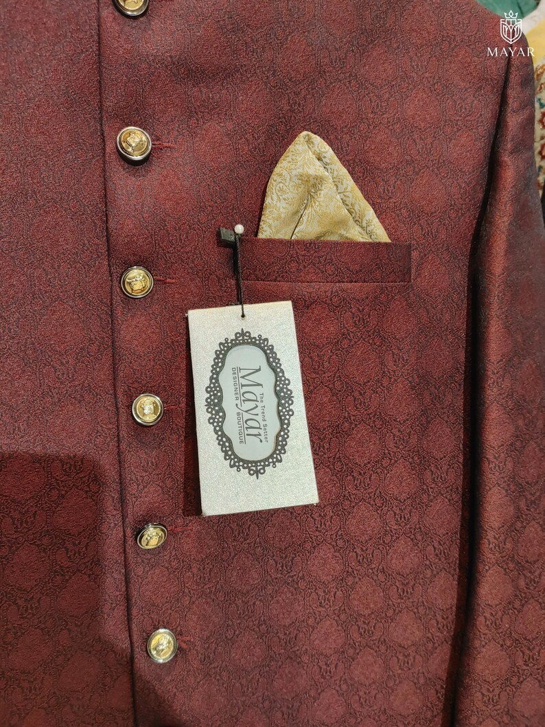 May include: A deep maroon jacket with a subtle repeating geometric pattern, featuring gold-toned buttons and a gold pocket square. A rectangular white tag hangs from the pocket, displaying the text Mayar The Trend Setter Designer Boutique.