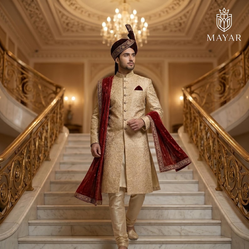 May include: A gold sherwani with intricate tonal embroidery, featuring a high collar, front button closure, and a pocket square. A deep maroon velvet turban and a matching maroon shawl with gold embroidered borders complete the ensemble.