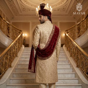 May include: A gold sherwani featuring intricate tonal embroidery, paired with a deep red velvet turban and a matching red velvet shawl draped over the shoulder. The garment includes a high collar and long sleeves, showcasing a detailed pattern.
